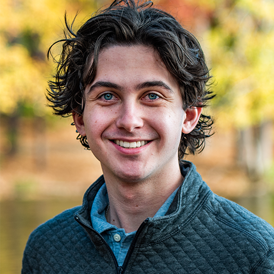 Luke Anderson in a blue shirt with a black jacket stands against yellow foliage in the background.