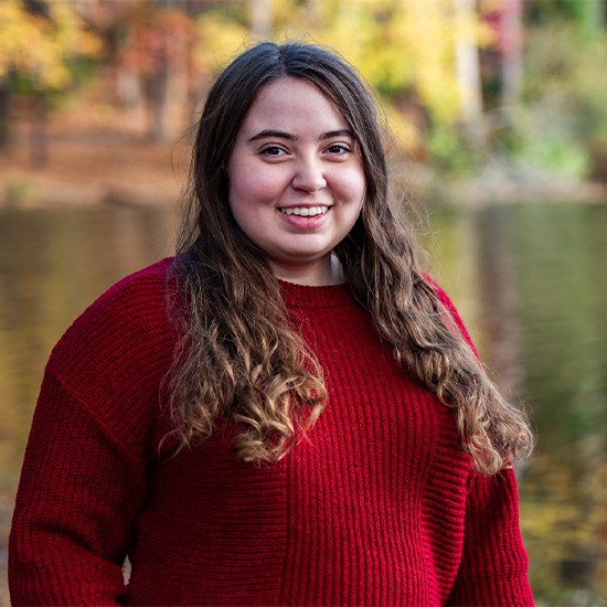 Alex Barr in a deep red sweater, stands against a lake with fall-colored foliage bei=hind them.