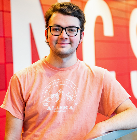 Tyler Negley in an orange t-shirt stands in front of a red wall with NC State written on it in big white letters.
