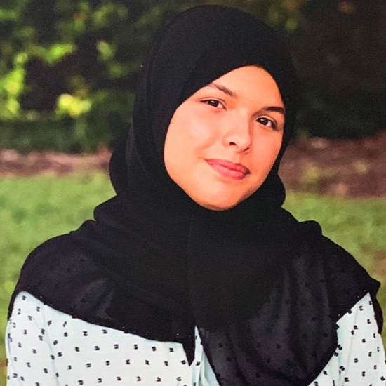 Eyman Sakr, in a black hijab with a black and white polka dot top, poses in front of green foliage.