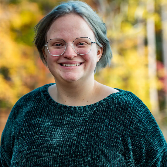 Theo Stewart, they/them, stands amongst yellow foliage in a green blouse.